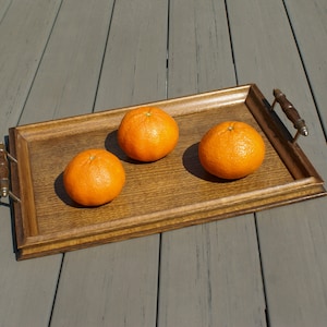 Könnte beinhalten: Ein rechteckiges Serviertablett aus Holz mit drei Orangen. Das Tablett hat eine dunkle Holzoberfläche und verzierte Griffe. Die Orangen sind leuchtend orange. Das Tablett steht auf einer grauen Holzoberfläche.