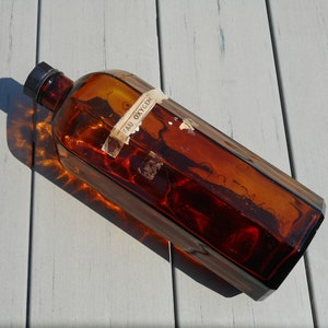 Op de afbeelding: Een vintage bruine glazen fles met een zwarte dop en een papieren label met de tekst "EAU OXYGEN". De fles heeft een rechthoekige vorm met afgeschuinde randen. De fles ligt op een lichtgrijze houten ondergrond.