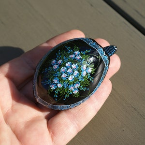 Könnte beinhalten: Eine schwarze, ovale Schmuckschatulle in Schildkrötenform. Der Deckel ist mit einem Blumenstrauß in Blau, Weiß und Rosa verziert. Die Ränder sind blau umrandet. Die Schatulle wird in einer Hand gehalten.