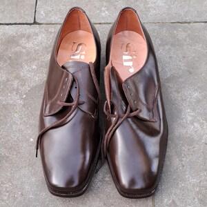 May include: A pair of brown leather dress shoes with laces. The shoes have a square toe and a polished finish. The word "Sir" is printed on the insole. The shoes are displayed on a gray surface.
