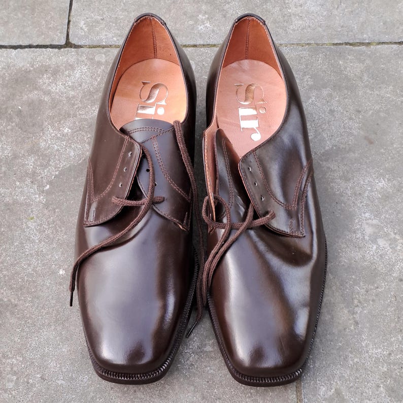 May include: A pair of brown leather dress shoes with laces. The shoes have a square toe and a polished finish. The word "Sir" is printed on the insole. The shoes are displayed on a gray surface.