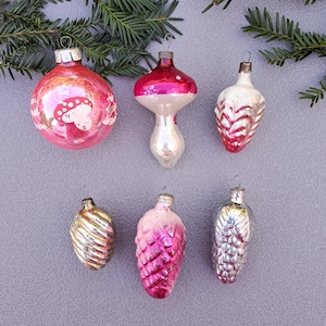 Op de afbeelding: Een verzameling vintage kerstversieringen in verschillende vormen en kleuren. Bevat een roze paddenstoelvormige ornament, een rood-witte druppel en verschillende dennenappelvormige ornamenten. De ornamenten worden tentoongesteld op een grijze ondergrond met groenblijvende takken.