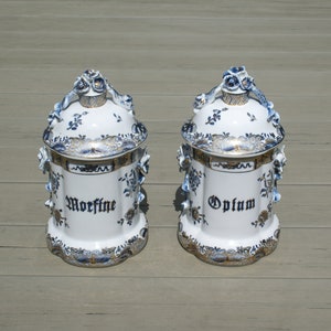 May include: Two white ceramic apothecary jars with blue floral designs and gold trim. One jar reads "Morphine" and the other reads "Opium" in dark blue script. The lids have floral handles.