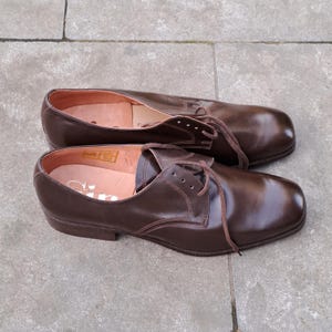 May include: A pair of brown leather dress shoes with laces. The shoes have a classic design with a rounded toe and a low heel. The shoes are placed on a gray stone surface.