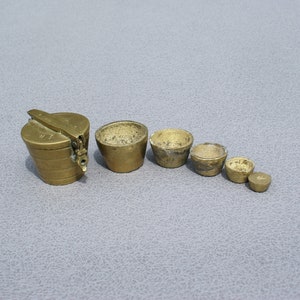 May include: A set of antique brass weights and a small container. The weights are in various sizes and shapes, including small, round, and cup-shaped. The container is cylindrical with a hinged lid. The items are arranged on a grey surface.