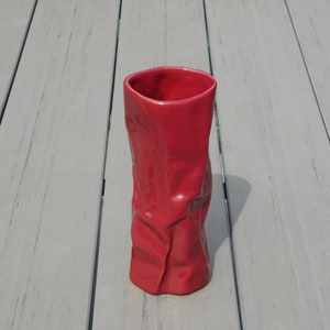 May include: A tall, red ceramic vase with a crumpled paper bag design. The vase has a cylindrical shape with a wide opening and a textured, uneven surface. The vase is approximately 8 inches tall.