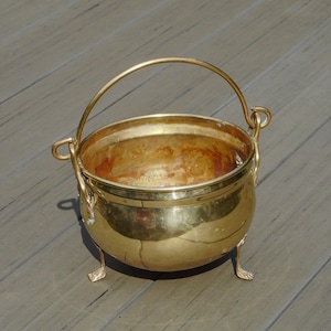 Op de afbeelding: Een glanzende, goudkleurige messing ketel met een gebogen handvat en drie kleine voetjes. De ketel heeft een ronde body en een brede opening. Het handvat is met decoratieve lussen aan de zijkanten bevestigd.