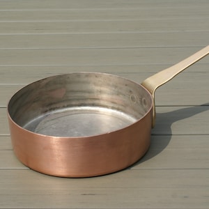 May include: A copper-colored saucepan with a long, brass-colored handle. The interior of the pan is silver-colored. The handle has a loop at the end for hanging. The pan is sitting on a gray wooden surface.