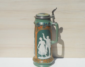 May include: Green ceramic beer stein with a hinged metal lid. The stein features a painted design of a dancing couple in white relief against a green background. The handle and base have an orange and gold trim.