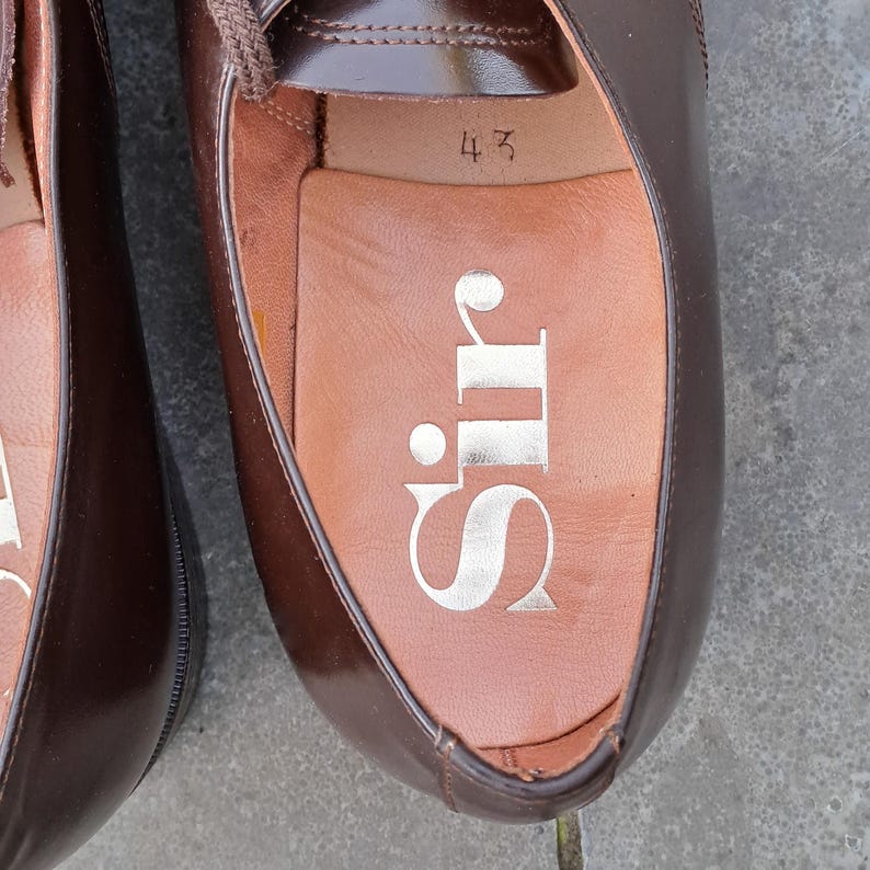 May include: Close-up of a pair of brown leather dress shoes. The interior of one shoe is visible, displaying the word "Sir" in gold lettering and the number "43". The shoes have laces and a glossy finish.