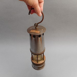 May include: Vintage metal miner's lamp with a cylindrical body, a copper-colored top and base, and a curved handle. The lamp has a glass section to view the light source and several ventilation holes. The lamp is held by a hand.