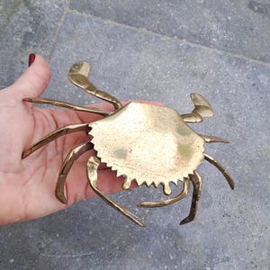May include: A brass crab sculpture with a textured surface and intricate details. The crab has multiple legs and claws, and a jagged edge around its body. The sculpture is held in a hand, showcasing its size and design.