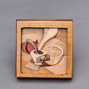 Könnte beinhalten: Holzintarsien-Kunstwerk, das ein Stillleben darstellt. Das Kunstwerk zeigt eine Tasse Kaffee, einen roten Apfel und eine Zitronenscheibe. Das Stück ist in einem hellbraunen Holzrahmen gerahmt, der die natürliche Holzmaserung zeigt.
