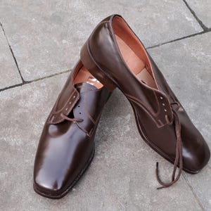 May include: A pair of brown leather dress shoes with a glossy finish. The shoes have a classic lace-up design and a square toe. The laces are brown, and the interior is a lighter brown color. The shoes are displayed on a gray surface.