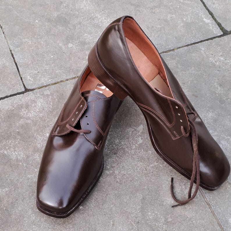 May include: A pair of brown leather dress shoes with a glossy finish. The shoes have a classic lace-up design and a square toe. The laces are brown, and the interior is a lighter brown color. The shoes are displayed on a gray surface.