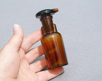 May include: A small, amber-colored glass bottle with a black stopper. The bottle has a rounded body and a narrow neck, held by a hand. The stopper has a small spout. The bottle is likely for liquids.