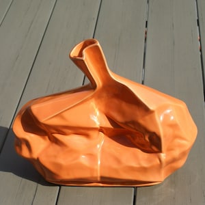 May include: An orange ceramic vase with a unique, crumpled design. The vase has a glossy finish and a folded top, creating an abstract and modern aesthetic. The vase is sitting on a gray wooden surface.