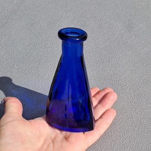Op de afbeelding: Een kobaltblauwe glazen fles met een uitlopende, driehoekige vorm. De fles heeft een afgeronde opening. Het glas heeft een diepe, rijke blauwe kleur en de fles lijkt vintage te zijn. Het wordt in een hand gehouden.