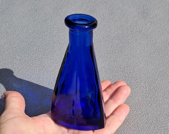 May include: A cobalt blue glass bottle with a flared, triangular shape. The bottle has a rounded opening and is held in a hand. The glass is a deep, rich blue color, and the bottle appears to be vintage.