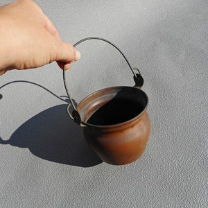 Op de afbeelding: Een kleine, koperkleurige ketel met een metalen handvat. De ketel is rond met een licht uitlopende rand. De handgreep is aan de zijkanten bevestigd. De ketel wordt tegen een grijze achtergrond gehouden.