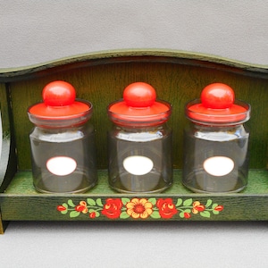 May include: A wooden spice rack with three clear glass jars, each topped with a bright red lid. The rack is painted green and decorated with a floral design along the bottom. The jars have blank white labels.
