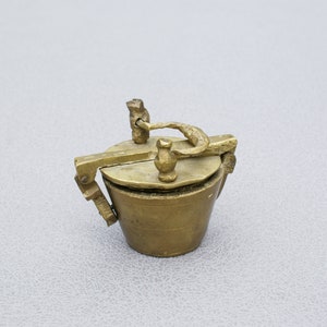 May include: A small, antique brass container with a hinged lid and a handle. The container has a cylindrical body and a circular lid with a small knob. The handle is curved and attached to the lid. The overall design is ornate and detailed.