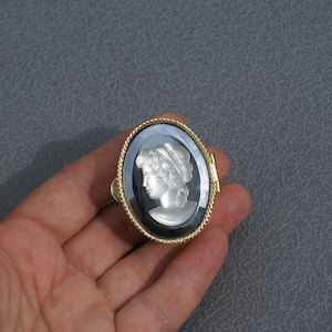 Op de afbeelding: Ovale camee-broche met een profiel van een vrouw met gedetailleerd haar en een hoofdbedekking. De camee is geplaatst tegen een zwarte achtergrond in een goudkleurige lijst met een touwachtige rand. De broche wordt in een hand gehouden.