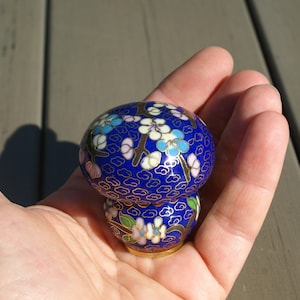 May include: A blue and gold cloisonné mushroom-shaped decorative object. The mushroom has a rounded top and a smaller base, both adorned with floral patterns in white, blue, and pink. The object is held in a hand.