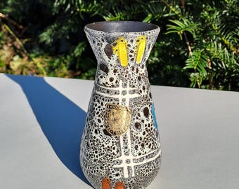 May include: A ceramic vase with a speckled black and white pattern, accented with yellow, blue, and orange details. The vase has a flared top and a gold emblem with text. A shadow is cast to the left.
