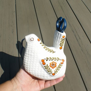 Könnte beinhalten: Weißer keramischer vogelförmiger Scherenhalter mit blauer Schere. Der Halter hat eine strukturierte Oberfläche und ist mit orangefarbenen und grünen floralen Designs verziert. Das Auge des Vogels ist ein einfacher schwarzer Punkt.