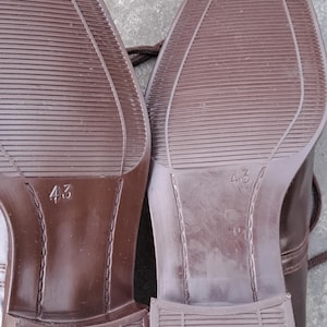 May include: Close-up of the soles of two brown leather shoes. The soles have a textured pattern and the number "43" is visible on each sole. The shoes appear to be new.