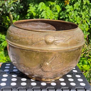 May include: An antique brass bowl with embossed floral and ribbon patterns. The bowl has a wide rim and a textured surface. The bowl is sitting on a black and white surface. The bowl's diameter is approximately 25 cm.