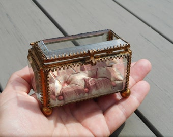 Könnte beinhalten: Kleine, rechteckige Glasschatulle mit Klappdeckel und messingfarbenem Metallbesatz. Der Innenraum zeigt ein rosa, getuftetes Kissen. Die Schatulle wird in einer Hand gehalten und zeigt ihr zartes Design und ihre Vintage-Ästhetik.