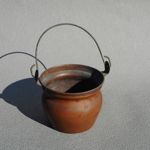 May include: A small, antique copper pot with a curved metal handle. The pot has a rounded body and a slightly flared rim. The copper has a warm, reddish-brown hue with some areas of green oxidation.