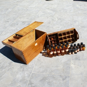 Op de afbeelding: Een vintage houten apothekerskist met een open deksel, een apart flessenrek en een verzameling kleine amberkleurige glazen flessen met etiketten. De kist en het rek zijn gemaakt van lichtbruin hout.