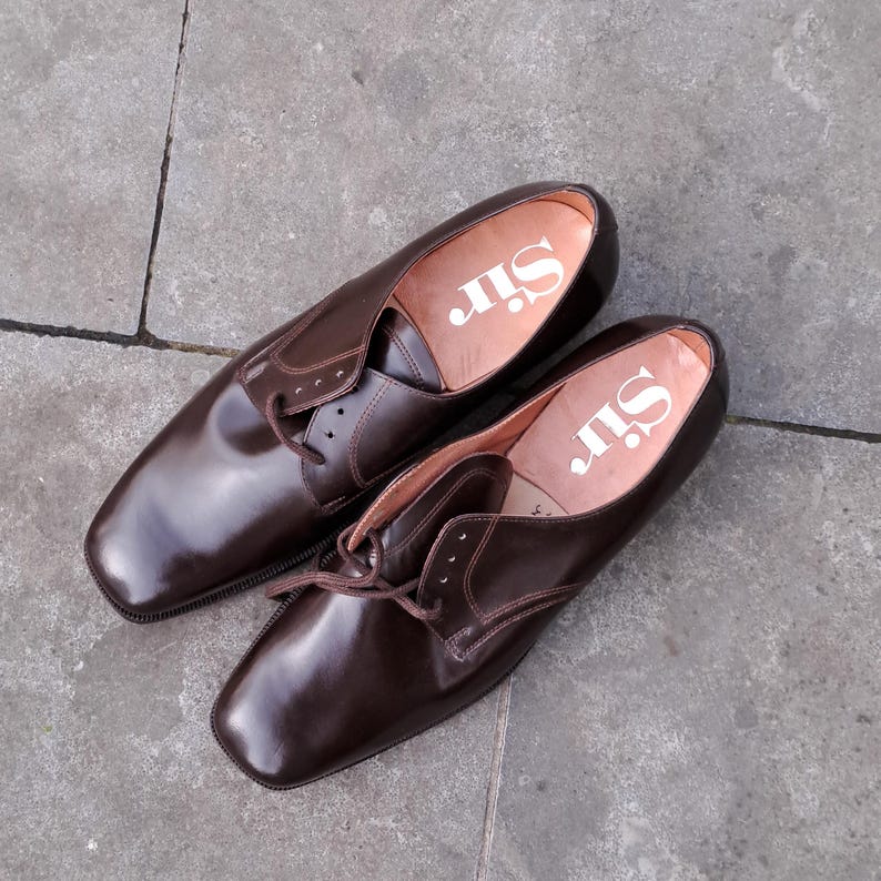 May include: A pair of brown leather dress shoes with a square toe and lace-up closure. The shoes have a glossy finish and the word "Sir" is printed in white on the insole.