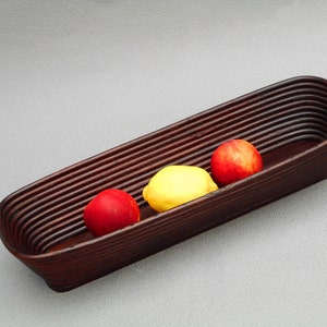 May include: A long, dark brown woven basket holds three pieces of fruit: two red nectarines and a yellow lemon. The basket has a curved, rectangular shape and is set against a gray background.