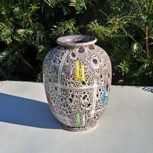 Op de afbeelding: Een decoratieve keramische vaas met een gespikkeld bruin en wit patroon. De vaas heeft een rasterontwerp met kleurrijke accenten, waaronder gele, blauwe en groene verticale lijnen. De vaas heeft een brede body en een smalle hals.