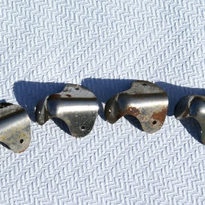 May include: Four vintage metal corner brackets with a curved design. The brackets are silver with areas of rust and small holes for mounting. They are arranged on a white textured surface.
