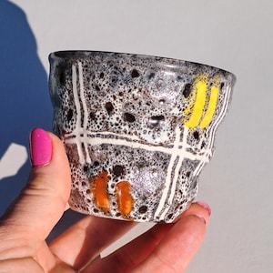 Könnte beinhalten: Eine handgefertigte Keramiktasse mit einer gesprenkelten schwarz-weißen Glasur. Die Tasse hat weiße Gitterlinien, gelbe Streifen und orangefarbene Akzente. Die Tasse wird in einer Hand gehalten, was ihre einzigartige, unregelmäßige Form hervorhebt.