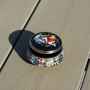 May include: A small, round, black trinket box with a gold rim and a lid featuring a painted scene of figures in blue and red. The box's sides are decorated with colorful figures. The box is open.