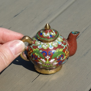 May include: A miniature, ornate teapot with a red base and lid, adorned with colorful floral patterns in green, blue, and white. The teapot features a gold handle, spout, and finial. A decorative item.