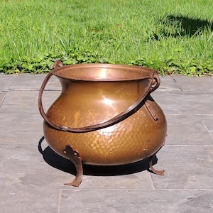 以下が含まれることがあります： ハンマー加工された質感、湾曲したハンドル、3つの装飾的な足が付いた銅製の大釜。大釜は丸みを帯びたボディと広い開口部を備えています。ハンドルは濃い色の金属でできています。大釜は石の表面に置かれています。
