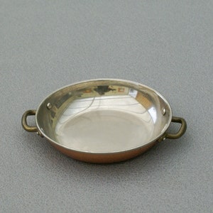 Peut inclure: Une petite poêle de couleur cuivre avec un intérieur argenté et deux poignées en laiton. La poêle est ronde et a une surface réfléchissante. La poêle est probablement utilisée pour cuisiner ou servir de petites portions de nourriture.
