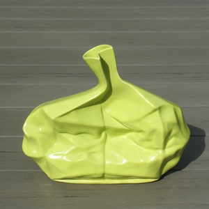 May include: A chartreuse-colored ceramic vase with a crumpled paper bag design. The vase has a unique, textured surface and a slender neck. The vase is sitting on a gray wooden surface.