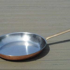 May include: A copper-bottomed frying pan with a stainless steel interior and a long, brass-colored handle. The pan is round and has a polished, reflective surface. The handle has a hole for hanging. Kitchenware.