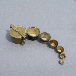 May include: A set of antique brass measuring cups and a lidded container. The cups are of varying sizes, arranged in a descending arc. The container has a hinged lid and a handle. The items are on a grey surface.