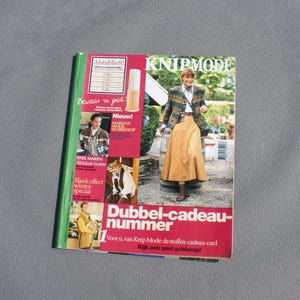 May include: A magazine cover featuring a woman in a plaid jacket and long skirt. The cover has the title "KNIPMODE" and various text blocks in Dutch. The magazine is open, revealing additional images and text.