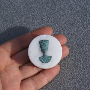 Op de afbeelding: Een witte, ronde decoratieve schijf met een turquoise buste van een Egyptische figuur. De buste is gecentreerd op het witte oppervlak. Het stuk wordt in een hand gehouden.