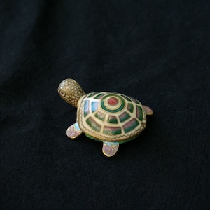 Könnte beinhalten: Eine dekorative Schildkrötenfigur mit detailliertem Schalenmuster. Die Schale hat eine cremefarbene Basis mit grünen, roten und rosa Akzenten, die goldfarben umrandet sind. Kopf und Beine der Schildkröte haben ein strukturiertes, gesprenkeltes Aussehen. Auf dunklem Hintergrund.